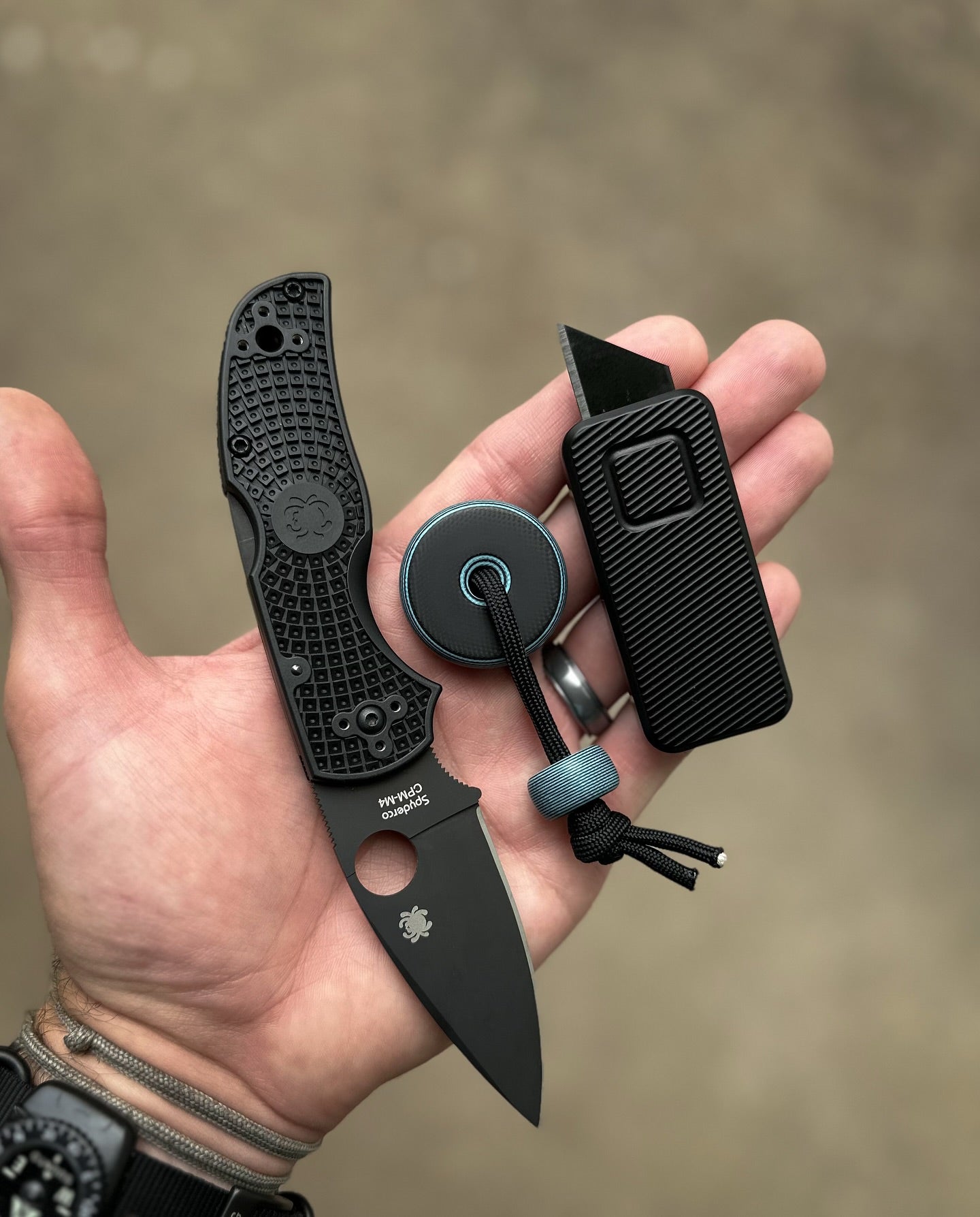 Zero knife in black with a larger black pocket knife and bead on a string, all displayed on an open hand with flat palm facing up