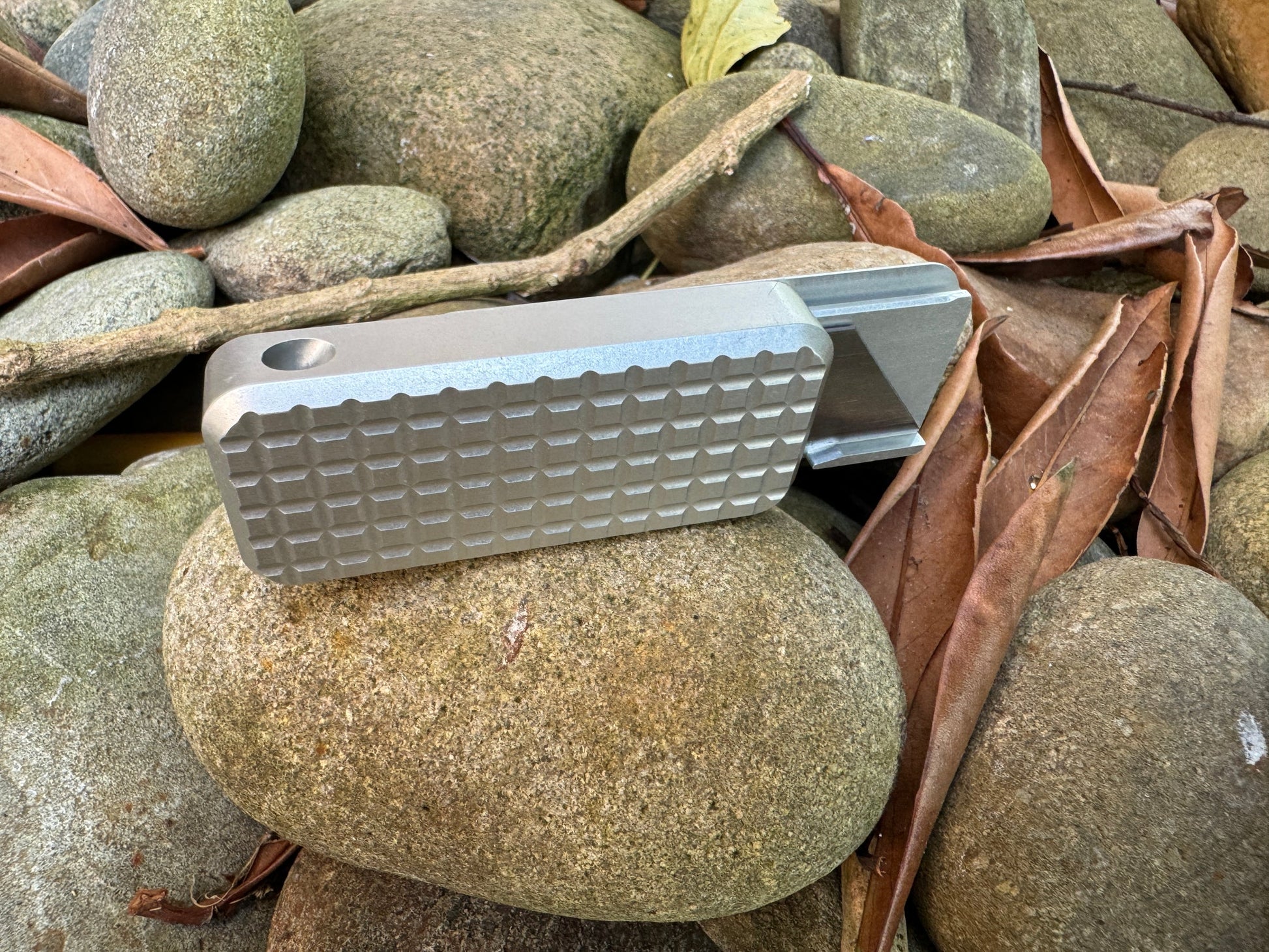 Back side of Rivery Zero knife showing lanyard hole with blade extended, sitting on a background of rocks and leaves