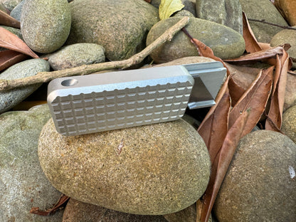 Back side of Rivery Zero knife showing lanyard hole with blade extended, sitting on a background of rocks and leaves