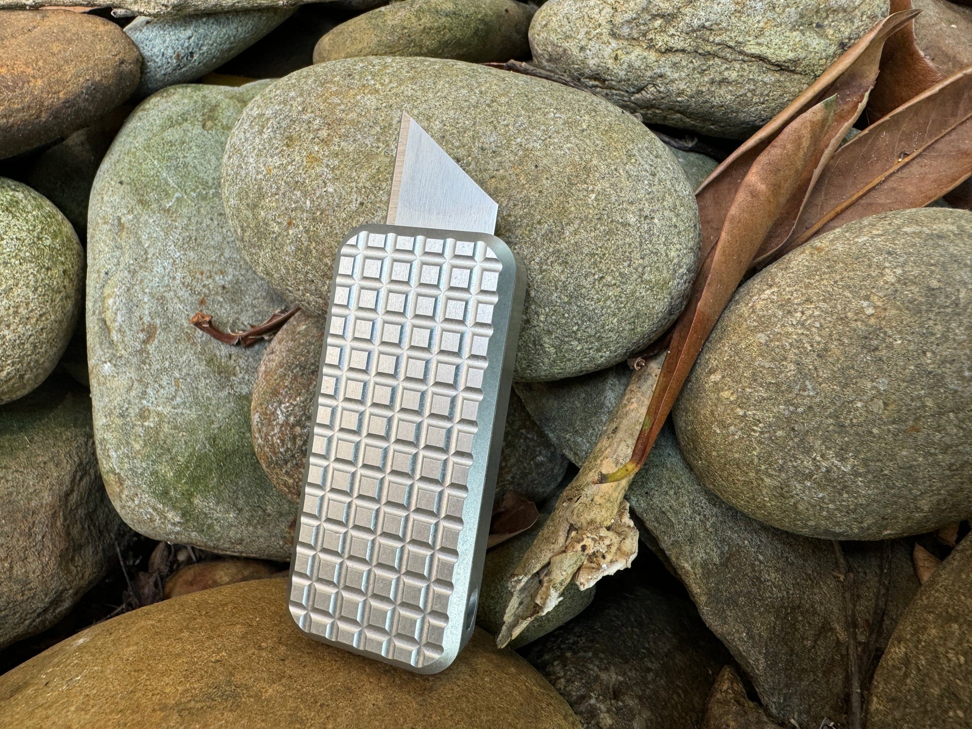 Rivery Zero knife with Grenade finish and blade extended, standing upright on a background of rocks and leaves