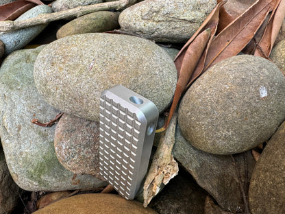 Bottom of Rivery Zero knife in Grenade finish showing lanyard hole, standing upright on a background of rocks and leaves