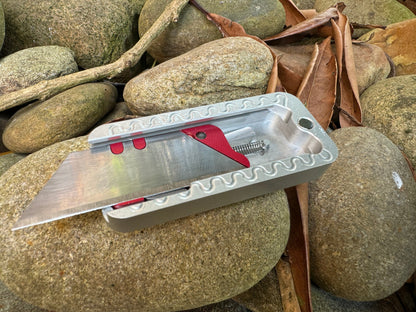 Rivery Zero knife with no lid, showing the internal utility blade, red carriage and spring, presented on a background of rocks, twigs and leaves