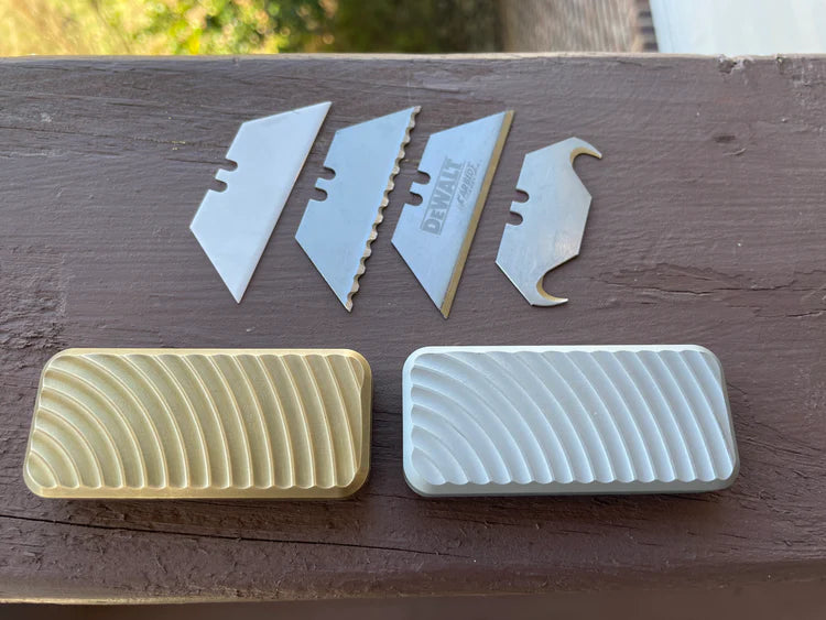 Four different types of utility blade, set above two Rivery Zero knives with Original finish, sitting on a brown painted piece of wood.